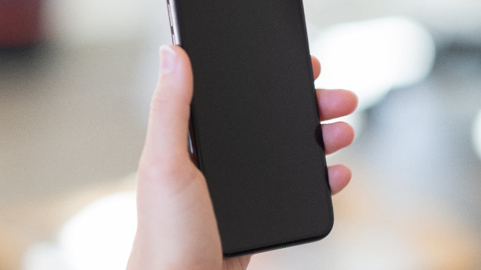 Mão segurando um celular de tela apagada, com o ambiente desfocado ao fundo